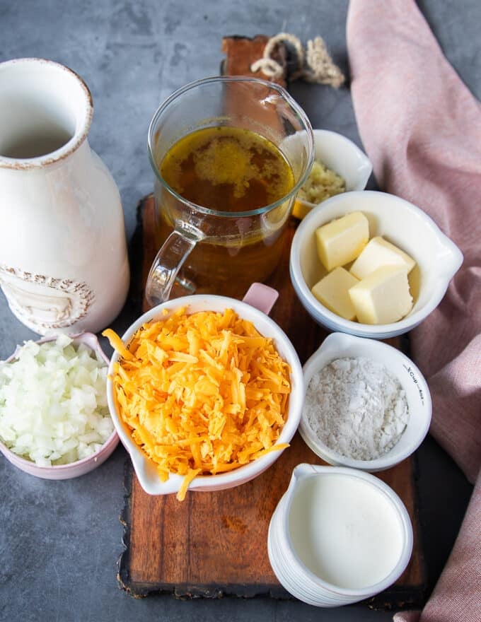 Baked potato soup ingredients in a bowl: a bowl of cheddar cheese, a bowl of flour, a bowl of diced onions, a bowl of butter, a jar of stock, a jar of milk, a jug of cream, a plate of minced garlic