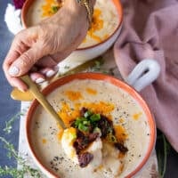 A hand holding a spoonful of baked potato soup toped with crumbles of sausage or bacon, green onions and cheddar cheese