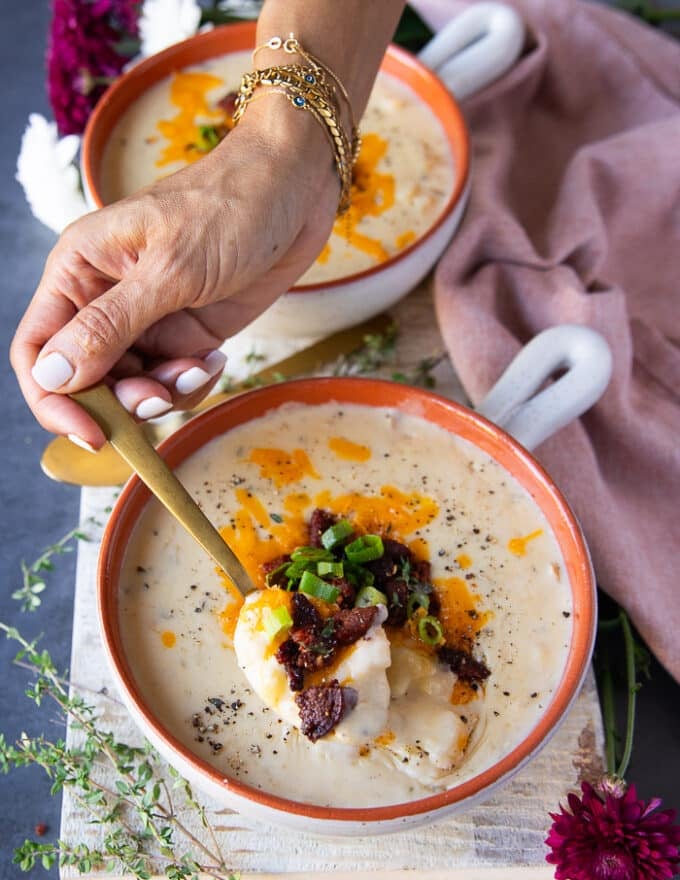 A hand holding a spoonful of baked potato soup toped with crumbles of sausage or bacon, green onions and cheddar cheese