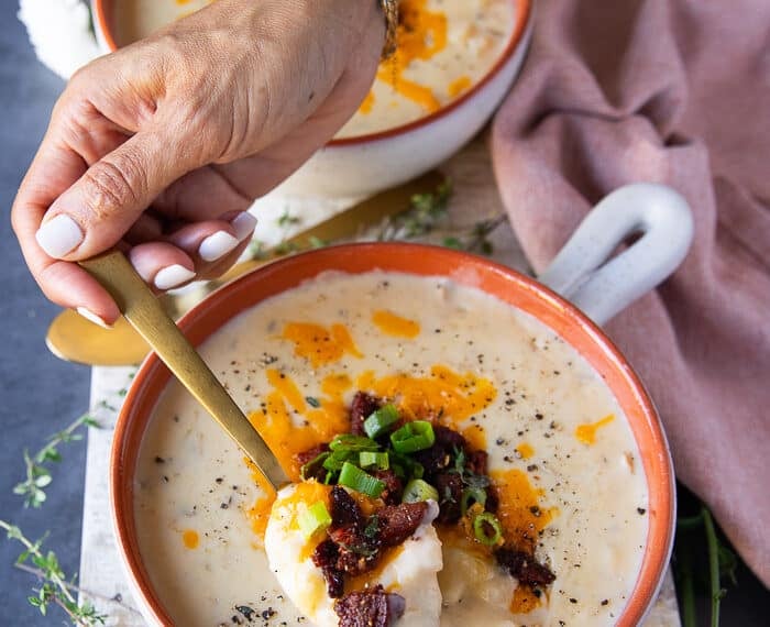 A hand holding a spoonful of baked potato soup toped with crumbles of sausage or bacon, green onions and cheddar cheese