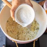 A hand adding in the flour into the onion and butter soup base in the pan