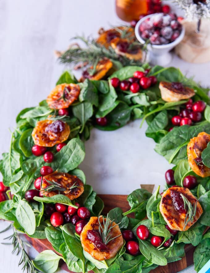 A wreath of spinach and cranberries on a white marble topped with cranberry brie bites and surrounded by frosted cranberries