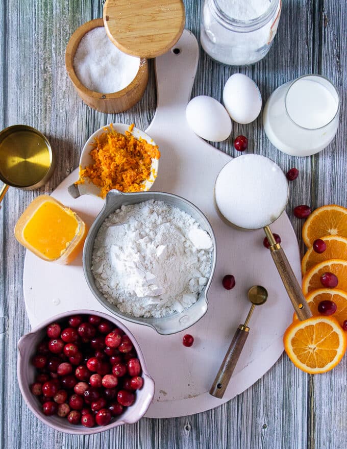 Ingredients to make cranberry orange muffins on a board including two eggs, a bowl of flour, a jar of baking powder, a bowl of oil, a bowl of orange zest, a cup of orange juice, a cup of milk, a bowl of fresh cranberries and a bowl of sugar