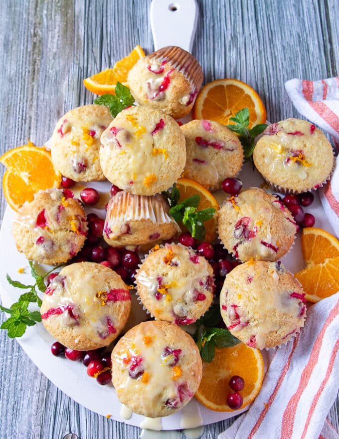 A final board of orange cranberry muffins glazed and ready to serve surrounded by an orange tea towel and some orange slices, some mint leaves and some fresh cranberries 