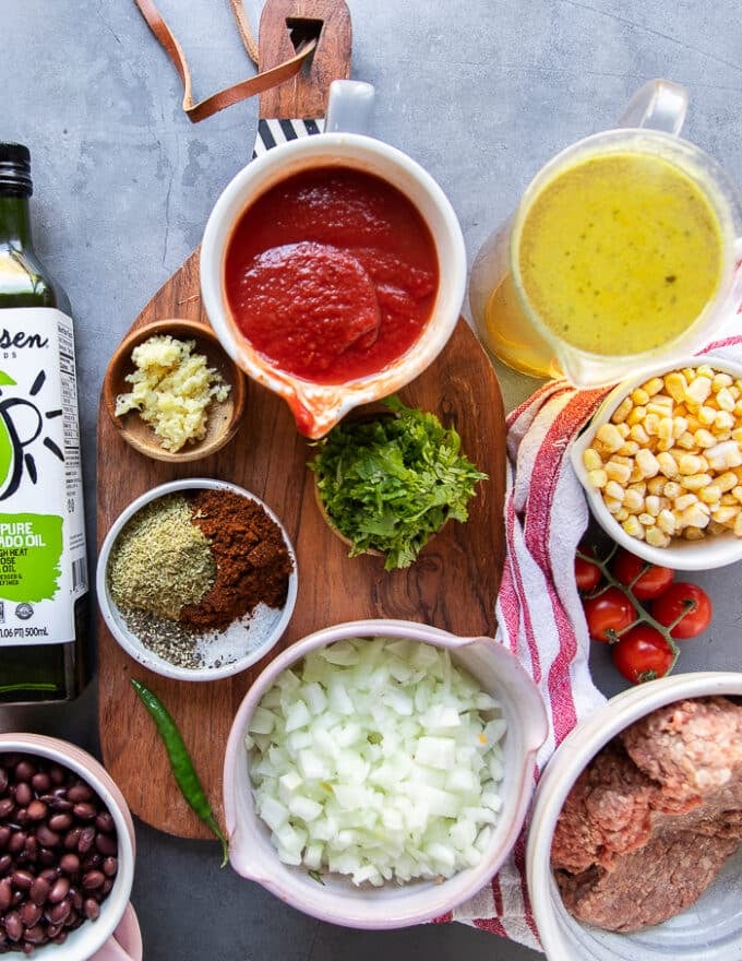 ingredients to make taco soup on a cooking board including: a jar of stock, a bowl of diced onions, another bowl of minced garlic, some chopped cilantro, a cup of tomatoes canned, a cup of frozen corn, a bowl of black beans, some ground beef, some oil, and a homemade taco spice blend and seasoning 