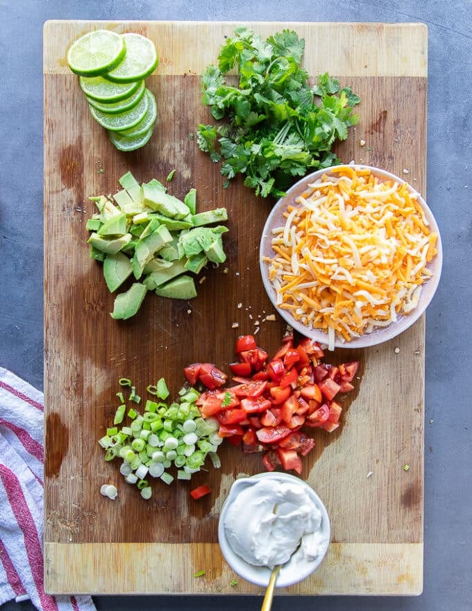 Topping ingredients for the taco soup including a bowl of shredded cheese, a cutting board with diced tomatoes, diced avocados, some chopped cilantro, some sour cream, mined green onions and lime slices