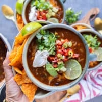 A. hand holding one bowl of taco soup showing a close up of the beans, tomato broth, corn, minced beef and the toppings like avocados, sour cream, cheese, a lime slice and cilantro