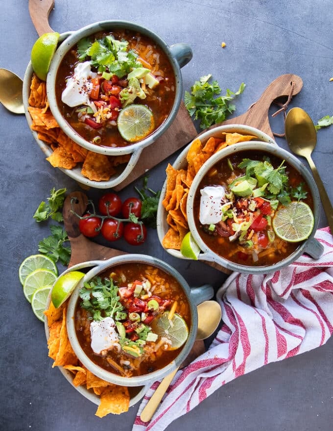 Three bowls of taco soup served up and garnished with different toppings like sour cream, cilantro, tomatoes, avocados, lime slices and chips