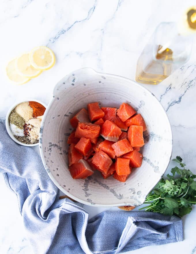 ingredients on a marbla top to make salmon bites including salmon cut up into bite size in a bowl, a plate of spice blend to season the salmon, some olive oil in a bottle