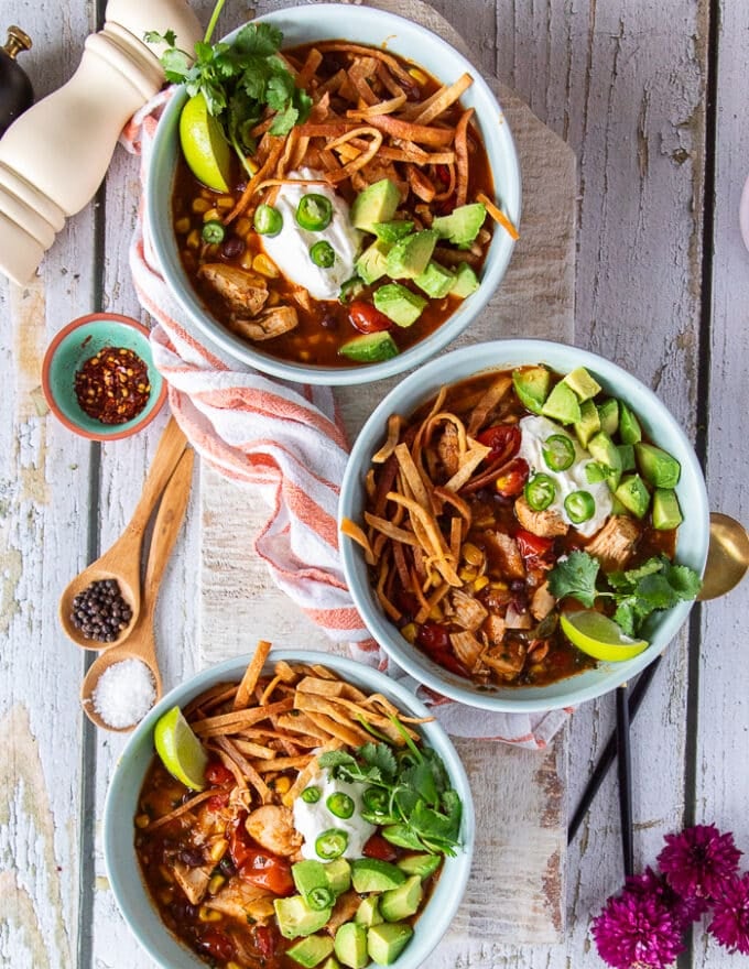 Three bowls of chicken tortilla soups on a wooden table loaded with tortilla chips, avocados, sour cream and green onions and surrounded by a kitchen towel