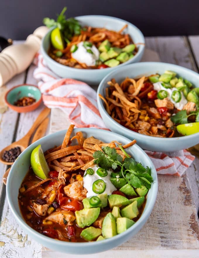close up of chicken tortilla soup in a bowl with sour cream, tortilla strips, jalapeno, avocados on top and lime wedges