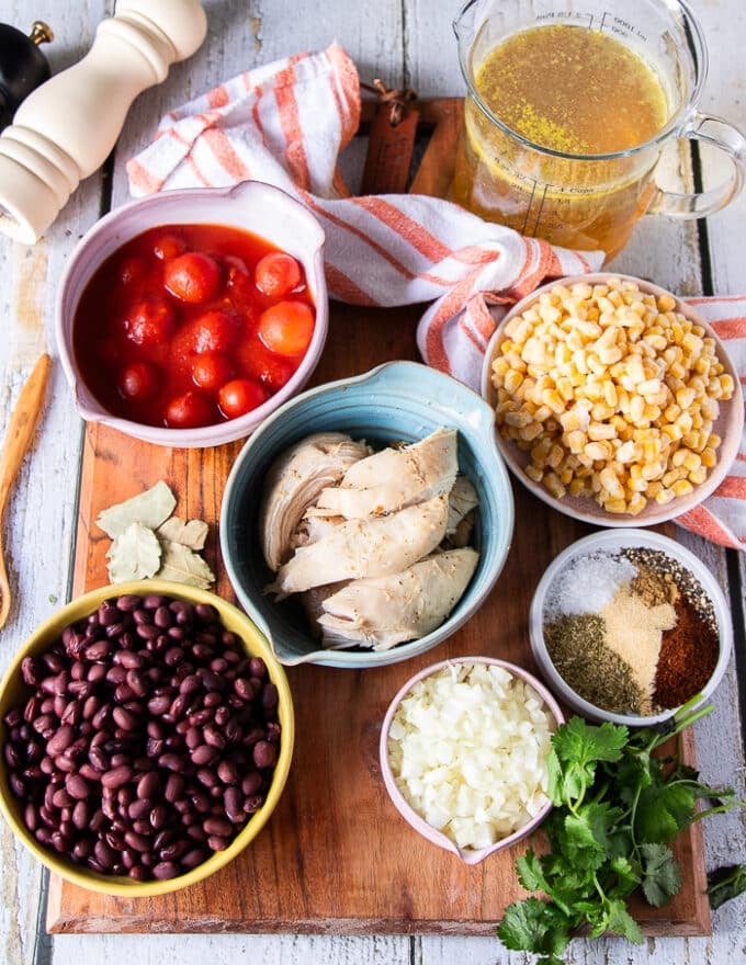 Ingredients for the chicken tortilla soup including the cooked chicken, some black beans in a bowl, some corn in a bowl, some canned tomatoes in a bowl, cilantro on a plate, some tortillas, a spice mix on a plate, some minced onions and garlic in smaller bowl and a jar of stock