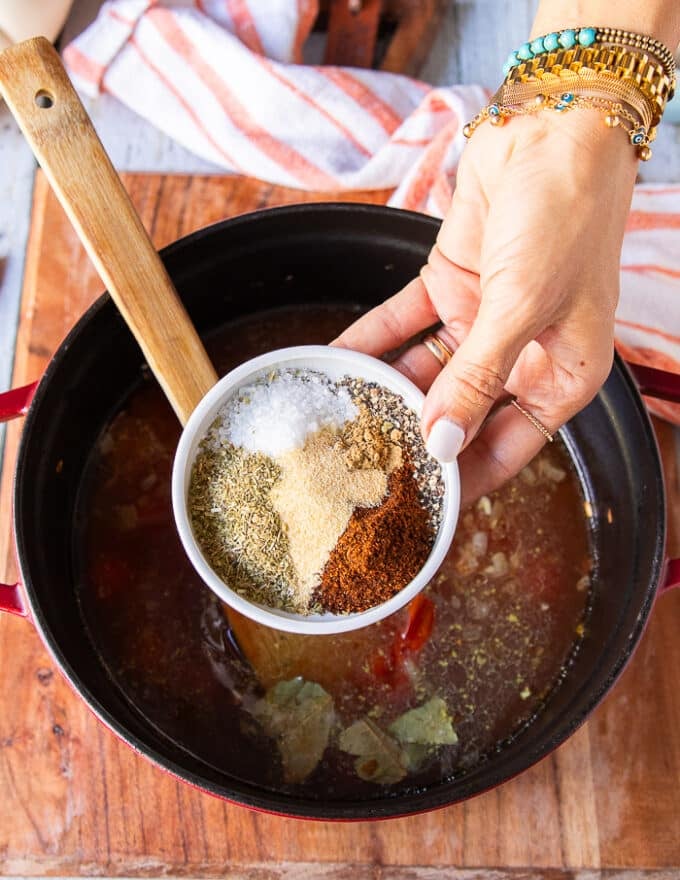 A hand holding the plate of different spices like the oregano, garlic powder, cumin, chilli powder, salt and pepper and adding it into the pot of soup