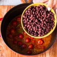A hand holding in the bowl of black beans and adding it into the soup pot