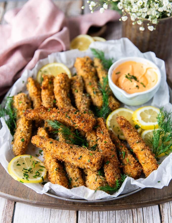 A plate of fish fingers out of the oven arranged on a plate surrounded by lemon slices, fresh dill and a bowl of dipping sauce