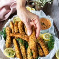 A hand holding one crunchy golden fish finger over a plate of fish fingers and some lemon slices