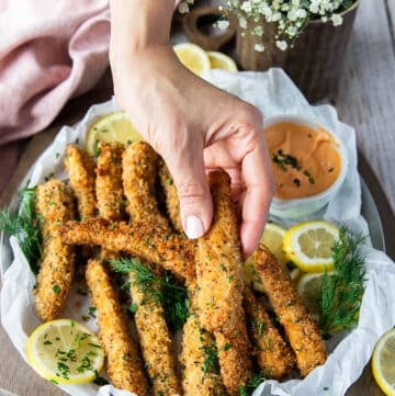 A hand holding one crunchy golden fish finger over a plate of fish fingers and some lemon slices