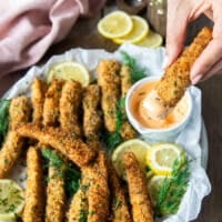 A hand dipping a fish fingers in a bowl of spicy mayo and tartare sauce