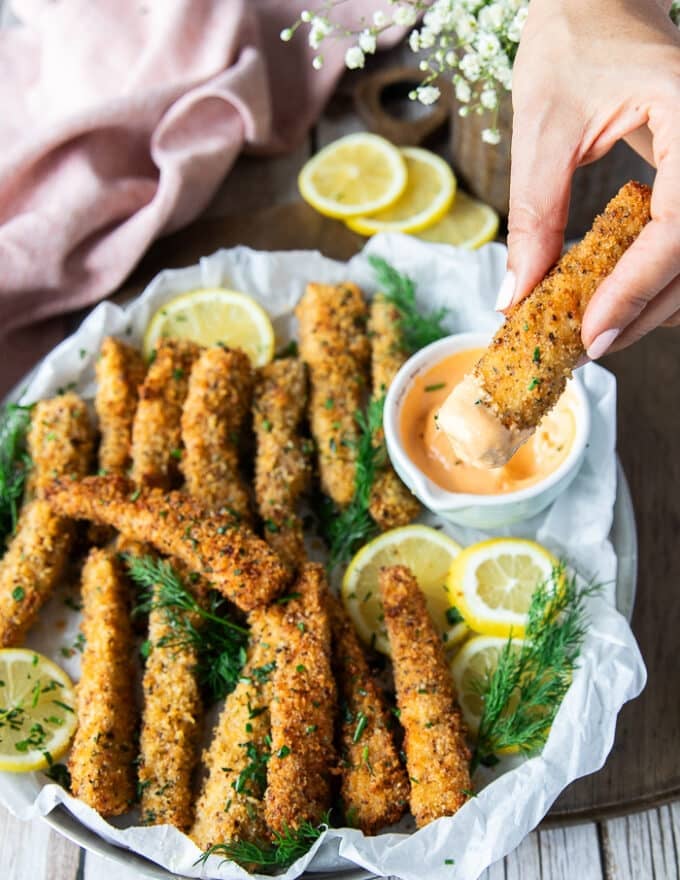 A hand dipping a fish fingers in a bowl of spicy mayo and tartare sauce 