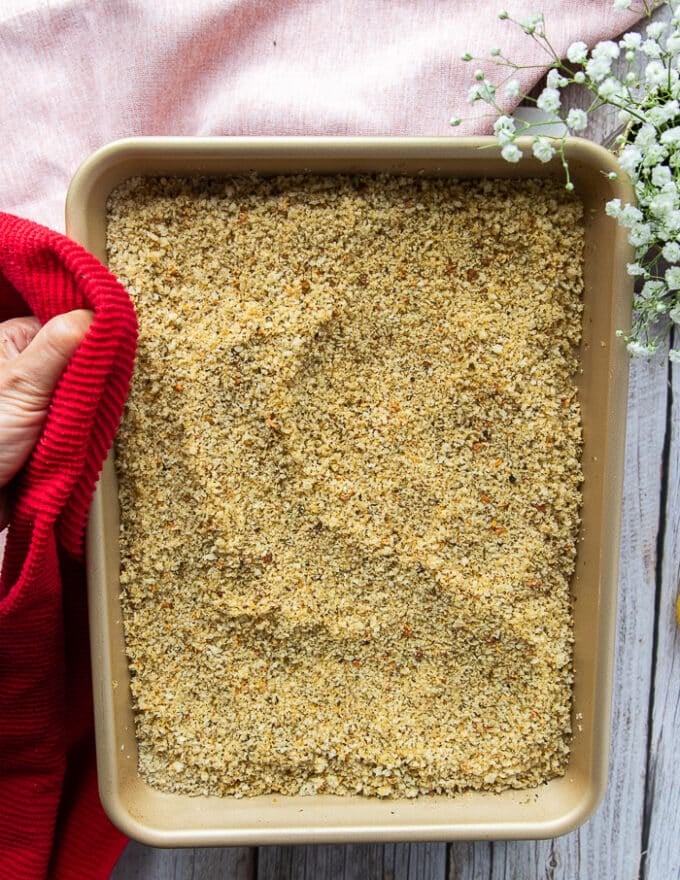 A hand holding a sheet pan of the toasted panko bread crumbs mixture 