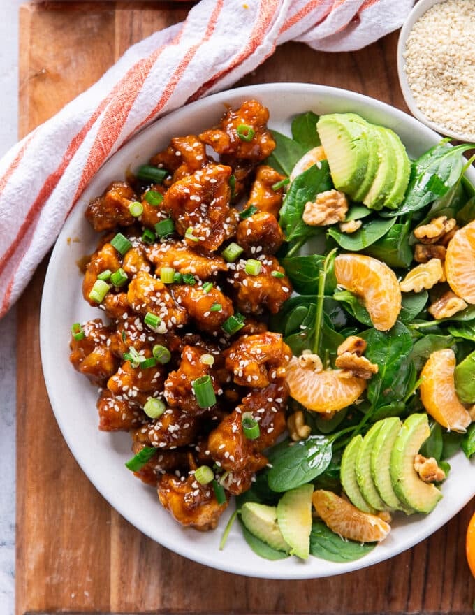 close up of a serving of homemade orange chicken on a plate showing the sticky orange chicken perfectly cooked and garnished with sesame and green onions along side some avocado salad 