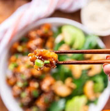 two chopsticks holding up a piece of orange chicken closeup showing the sauce and texture of the chicken with sesame seed stuck on it