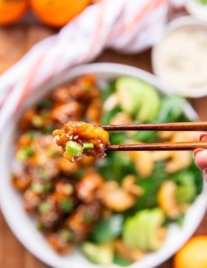 two chopsticks holding up a piece of orange chicken closeup showing the sauce and texture of the chicken with sesame seed stuck on it