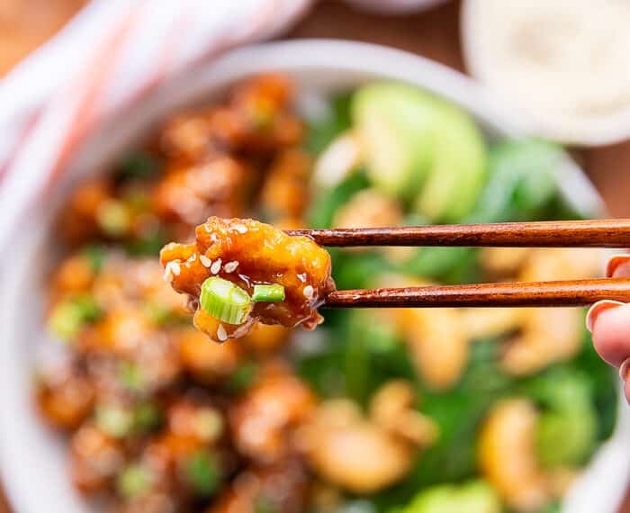 two chopsticks holding up a piece of orange chicken closeup showing the sauce and texture of the chicken with sesame seed stuck on it