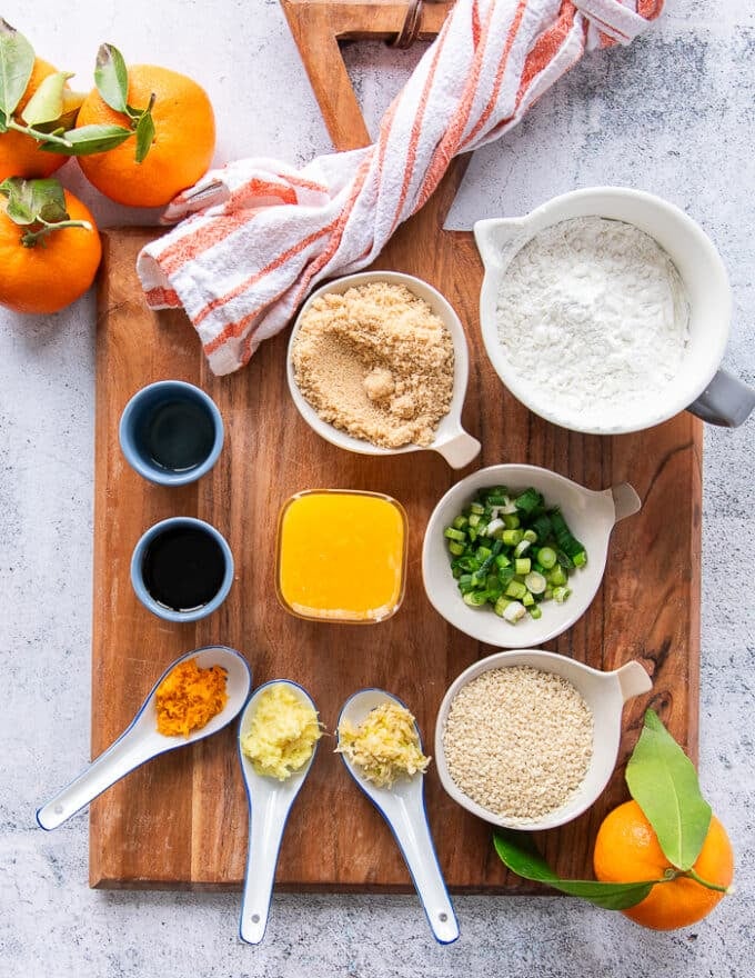 orange chicken sauce ingredients on a cutting board including cornstarch and water mixed in a bowl, orange juice in a cup, orange zest on the board, grated garlic in a bowl, grated ginger in a bowl. soy sauce in a bowl, rice vinegar in a bowl, brown sugar in a bowl and green onions and sesame seeds 