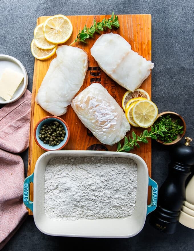 ingredients for pan seared cod recipe on a wooden board inluding a plate of cod fish, a plate of flour, some lemon juice in a bowl, lemon zest, butter in a bowl, fresh oregano on the board, capers in a bowl, stock in a cup, salt and pepper mills