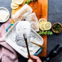 A hand holding a cod fish dusted lightly with flour and showing how lightly coated it is above the flour plate