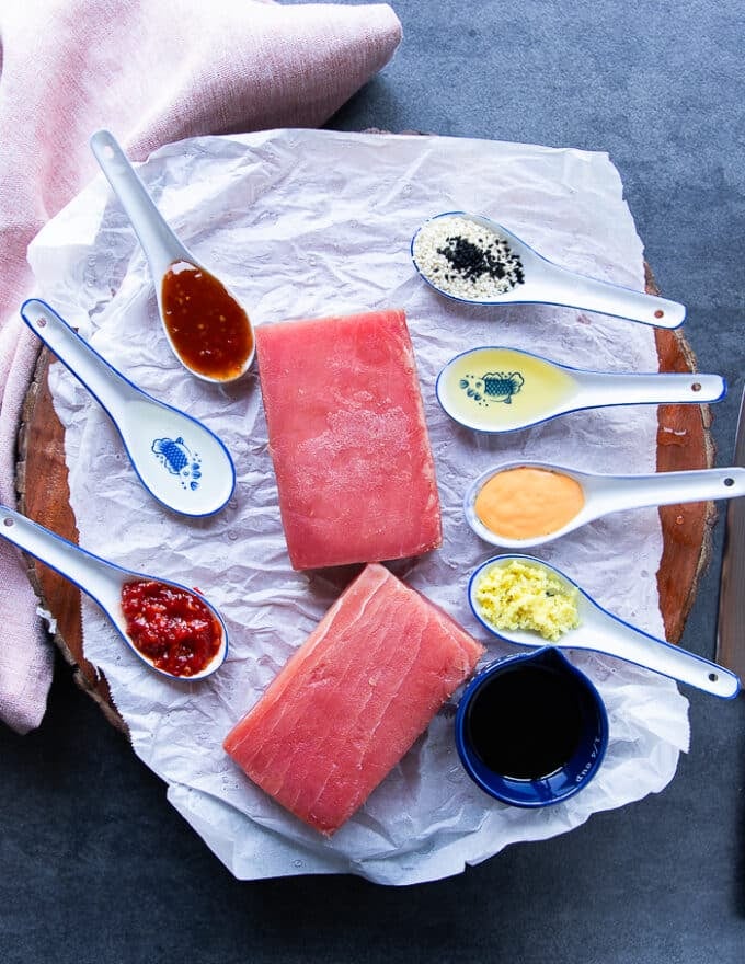 ingredients for poke bowl on a cutting board including saku tuna, small bowls of fresh ginger, sriracha, rice vinegar, chilli sauce, soy sauce, sesame seeds, spicy mayo