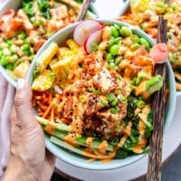 A hand holding a poke bowl very close with chopsticks. The poke bowl includes sushi tuna, rice, carrots, edamame , seaweed salad, pickled ginger, cucumbers all drizzled with spicy mayo