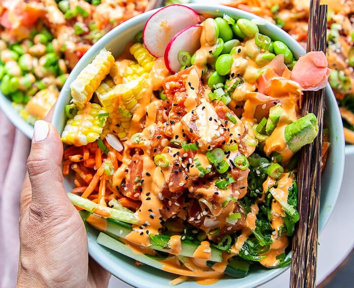 A hand holding a poke bowl very close with chopsticks. The poke bowl includes sushi tuna, rice, carrots, edamame , seaweed salad, pickled ginger, cucumbers all drizzled with spicy mayo