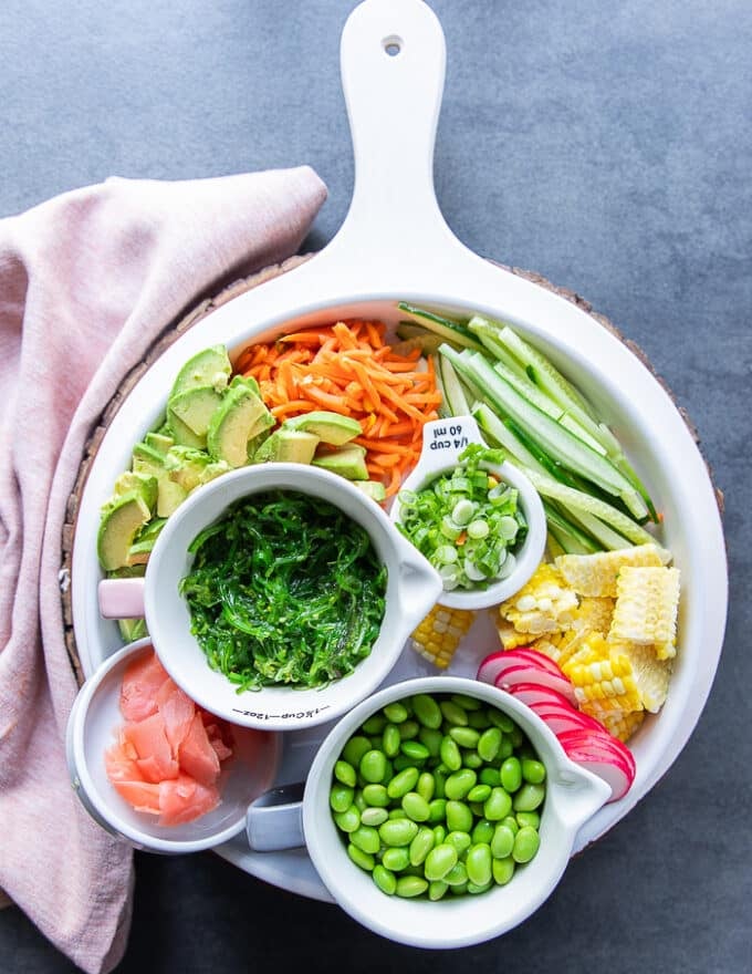 Veggies to assemble the poke bowls in small bowls. Bowls of edamame, pickled ginger, corn, seaweed salad, cucumbers sliced, carrot sticks, sliced avocados