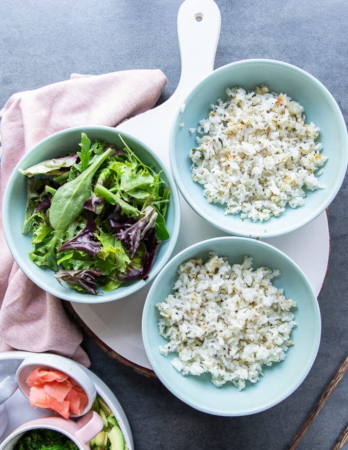Three bowls on a table, some bowls have sushi rice as a base, some bowls have salad as a base ready to assemble the bowls