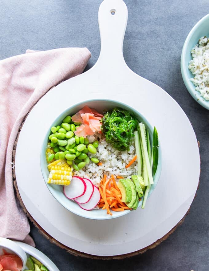 The veggies are layered around the bowl with sushi rice including some edamame, cucumbers, seaweed, carrots, pickled ginger, corn, radishes