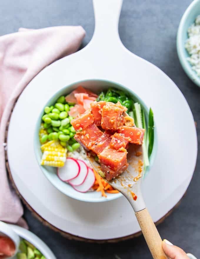 A hand holding a spatula and scooping in the diced chunks of seasoned tuna in to the center of the poke bowls