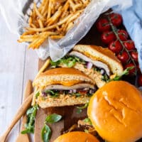 Three portobello mushroom burgers on a wooden plate with one of them cut in half showing the texture and filling of the burger and the portobello mushroom. The burgers are surrounded by a plate of french fries