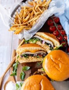 Three portobello mushroom burgers on a wooden plate with one of them cut in half showing the texture and filling of the burger and the portobello mushroom. The burgers are surrounded by a plate of french fries