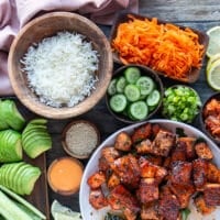 ingredients needed to make salmon bowl: cooked salmon bites in a bowl, coked rice in a bowl, shredded carrot on a plate, sliced avocados on a board, sliced cucumbers and green onions on a board, some spicy mayo, sesame seeds in a bowl, shredded seaweed on a bowl