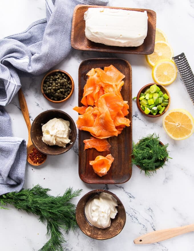 smoked salmon dip ingredients in a wooden board. a plate of smoked salmon, a bowl of cream cheese, some sour cream in a bowl mayonnaise in another bowl. capers in a small plate, green onions in a small plate, seasoning, some fresh dill in a bowl, and lemons cut into wedges and slices