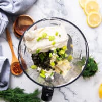 The process of making smoked salmon dip starts with cream cheese, sour cream and mayonnaise along with green onions and capers along with lemon zest and juice all added into a bowl of a food processor.