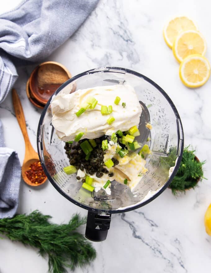 The process of making smoked salmon dip starts with cream cheese, sour cream and mayonnaise along with green onions and capers along with lemon zest and juice all added into a bowl of a food processor. 