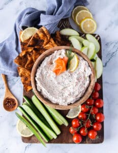 A bowl of smoked salmon dip on a wooden board surrounded by pita chips, cucumber, tomatoes and apples.