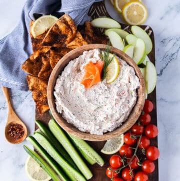 A bowl of smoked salmon dip on a wooden board surrounded by pita chips, cucumber, tomatoes and apples.