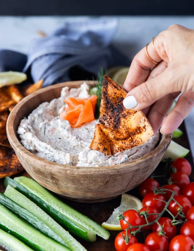 A hand holding a pita chip and dipping it right into the smoked salmon dip bowl showing the consistency of the dip