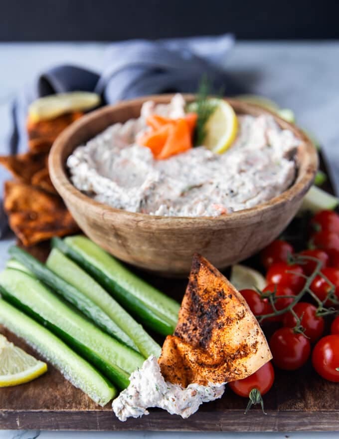 A side view close up of a bowl of smoked salmon dip with a topping of one smoked salmon curled up and a pita chip with some dip on it near the bowl