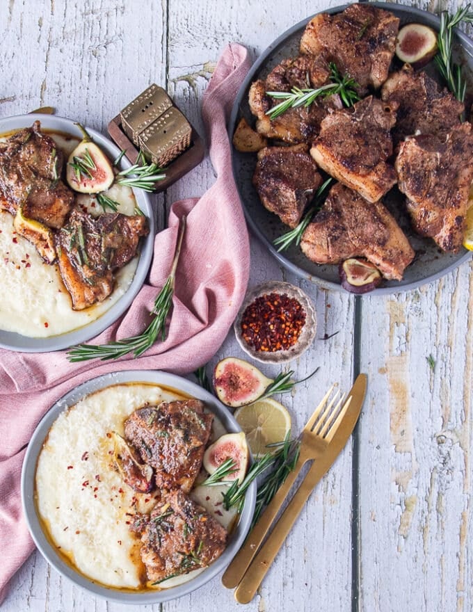 A table set up of a plate of cooked lamb loin chops on the side and then a few plates with mashed potatoes, some greens, some fresh figs, fresh rosemary and lemon slices for serving the lamb loin chops