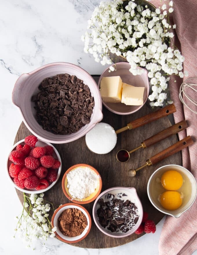 Ingredients to make molten lava cakes on a wooden board including chocolate chunks in a bowl, unsalted butter in a plate, some flour in a bowl, some cocoa powder in a bowl. some eggs in a bowl, some cream in a bowl and some sugar in a measuring cup